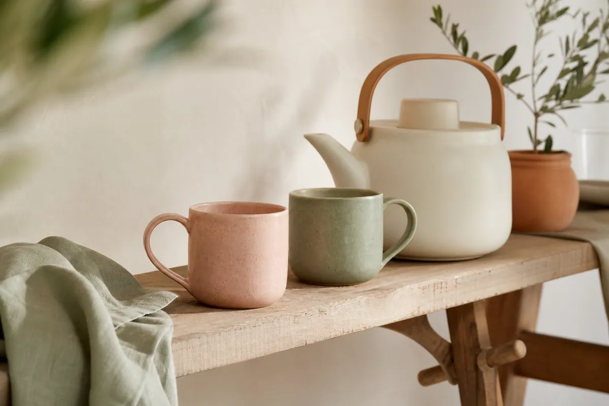 Blush pink and sage green mugs on a bare timber bench beside a kettle